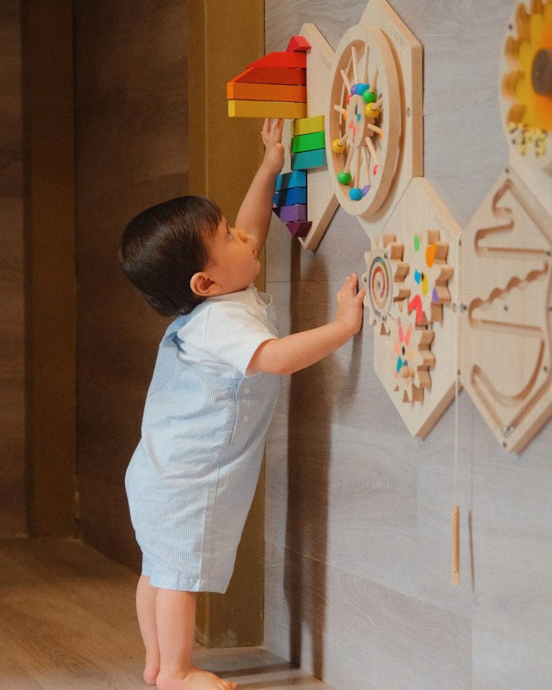 Um bebê de pé diante de um painel de brinquedos de madeira, com a mão no painel, de costas para a câmera.