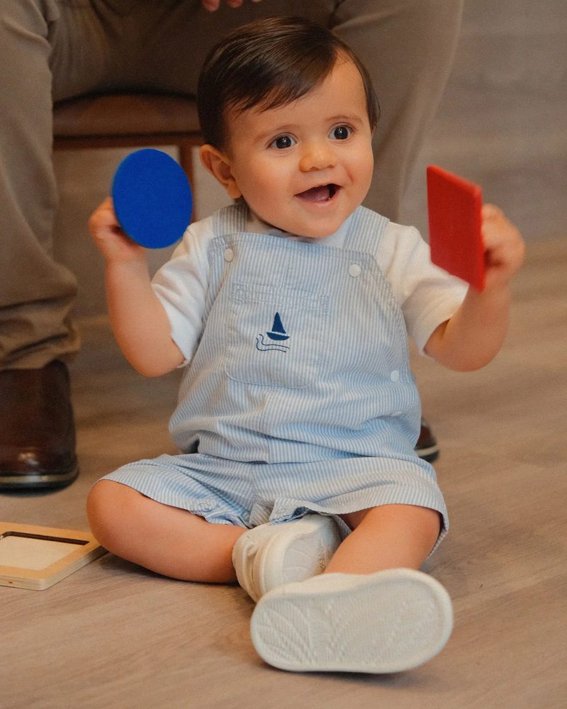 Um bebê sentado no chão, sorrindo, com uma plaquinha azul numa mão e uma vermelha na outra.