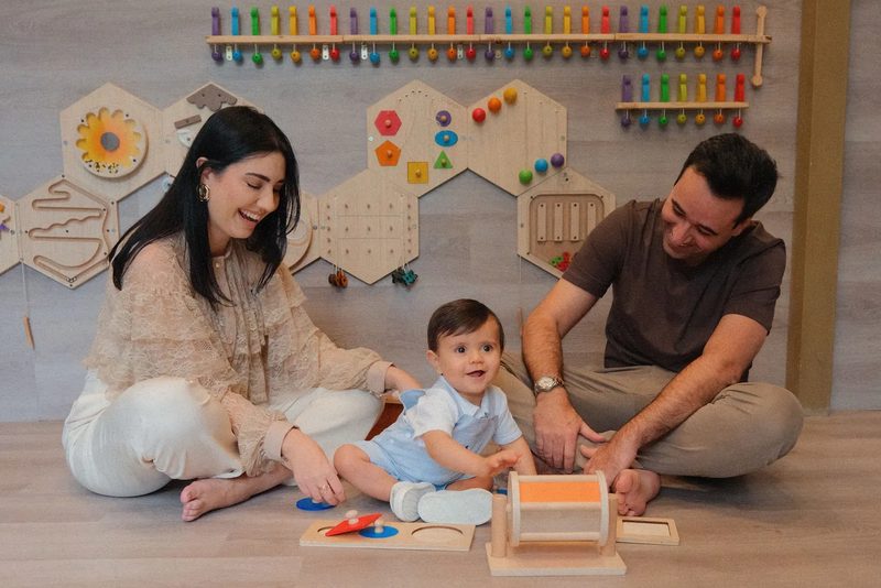 Tana, Rafael e o bebê sentados no chão, sorrindo e brincando com peças de madeira, diante de uma parede de brinquedos.