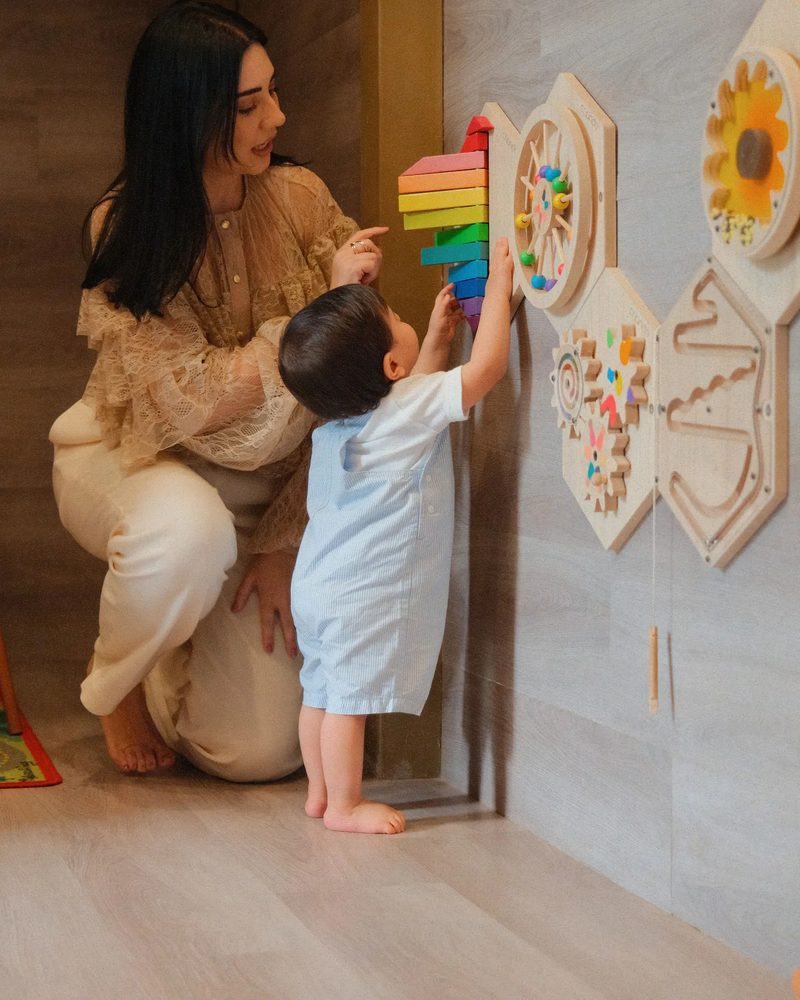 Tana agachada ao lado de um bebê de pé diante de um painel de brinquedos, observando enquanto ele alcança as peças.