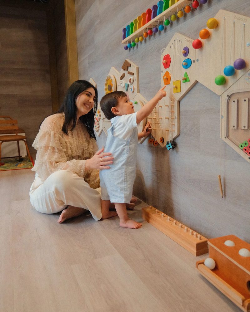 Tana sentada no chão, sorrindo, com a mão nas costas de um bebê de pé que aponta para um painel de brinquedos de madeira.